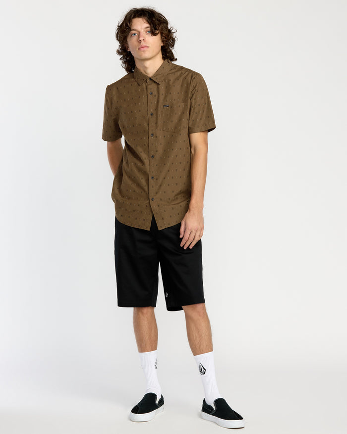 A person with curly hair wears a Volcom Men's Bankstone Short Sleeve Shirt in brown, classic fit, black knee-length shorts, white socks with a black logo, and black slip-on shoes, standing against a plain white background.