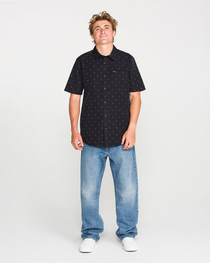 A person stands against a plain white background, wearing the Volcom Men's Bankstone Short Sleeve Shirt, featuring a small pattern, paired with light blue jeans and white sneakers. The classic fit shirt is crafted from soft cotton-poly fabric for all-day comfort and style.