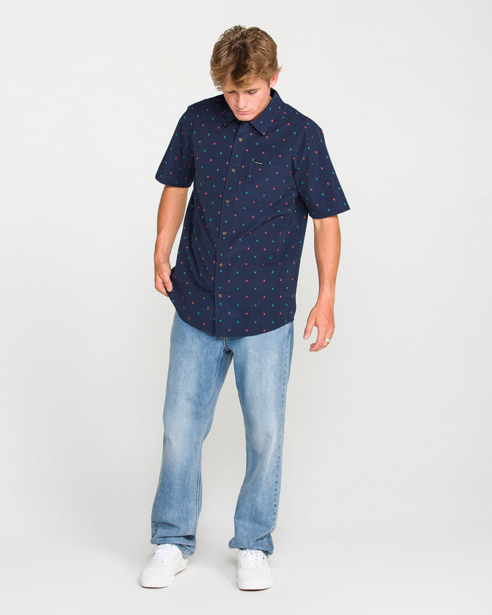 A young man wearing the Volcom Men's Bankstone Short Sleeve Shirt in a classic fit with a navy dobby weave and red pattern, paired with light blue jeans and white sneakers, stands against a plain white background, looking down.