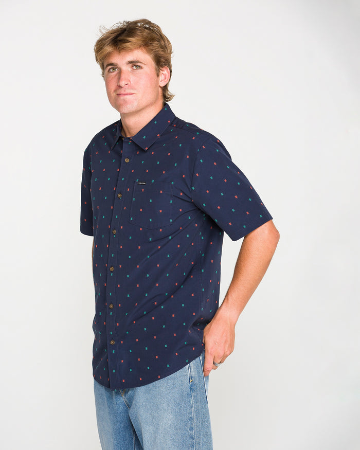 Wearing the Men's Bankstone Short Sleeve Shirt by Volcom in navy blue, featuring a classic fit and subtle dobby weave pattern, a person pairs it with light blue jeans and stands with one hand in their pocket against a plain white background.
