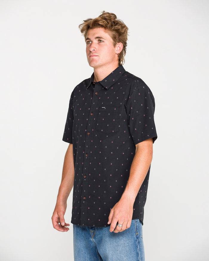 A man is wearing the Volcom Men's Bankstone Short Sleeve Shirt—a classic fit button-up with a subtle dotted pattern—paired with light blue jeans, standing facing left against a plain white background. This cotton-poly shirt from Volcom provides both comfort and style for everyday wear.