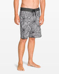 A man stands barefoot against a plain white background, wearing Volcom Men's Barnacle Scallop Stoney black and white patterned boardshorts featuring the Stoney Cinch Fly.