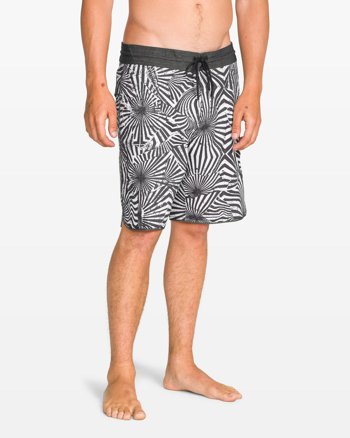 A man stands barefoot against a plain white background, wearing Volcom Men's Barnacle Scallop Stoney black and white patterned boardshorts featuring the Stoney Cinch Fly.