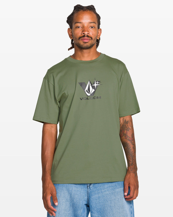 A man with dreadlocks wears the Volcom Men's Be Kind Rewind Short Sleeve Tee in olive green and light blue jeans, standing against a plain white background.