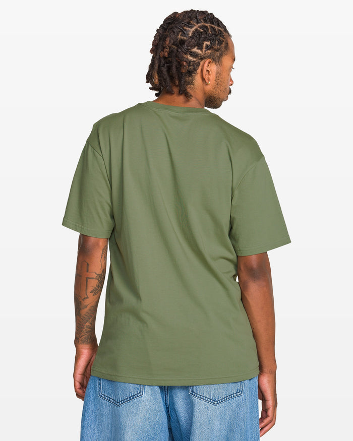 A man with short dreadlocks, dressed in Volcom’s Men's Be Kind Rewind Short Sleeve Tee and light blue jeans, stands with his back to the camera against a plain white background.