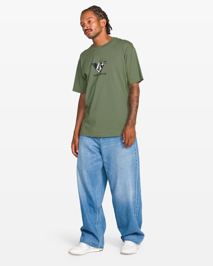 A man in a Volcom Men's Be Kind Rewind Short Sleeve Tee (classic fit, olive green), baggy light blue jeans, and white sneakers stands against a plain white background.