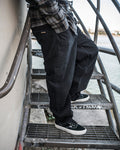 A person in a plaid shirt and Volcom Men’s Billow Denim Pants stands on outdoor metal stairs by the water, with graffiti visible on the railing and steps.