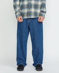 A person wearing a gray plaid shirt, Volcom Men's Billow Denim Pants in a loose fit, and black sneakers stands against a plain white background. The photo shows their body from the shoulders down.