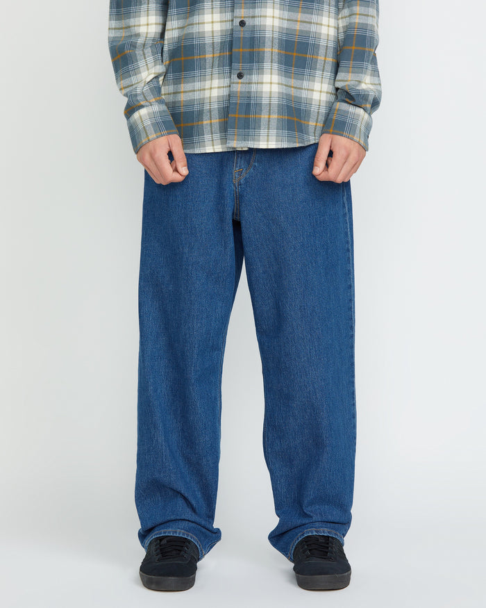A person wearing a gray plaid shirt, Volcom Men's Billow Denim Pants in a loose fit, and black sneakers stands against a plain white background. The photo shows their body from the shoulders down.