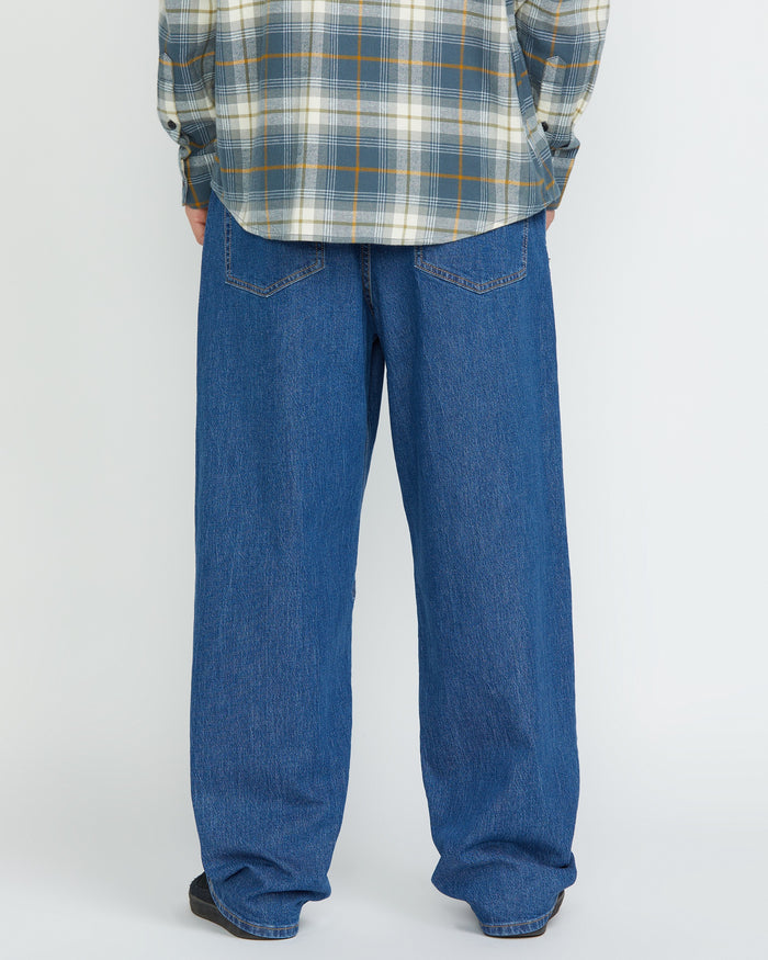 A person stands facing away, showing only the lower half, wearing Volcom Men's Billow Denim Pants and a gray plaid shirt against a plain light background.