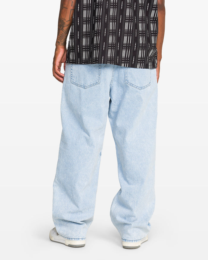 A person faces away, wearing a black and white patterned shirt, light blue Volcom Men's Billow Denim Pant made from recycled cotton, and white sneakers. The background is plain white.