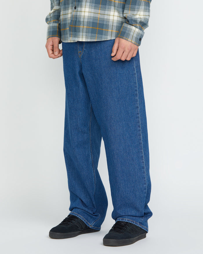 A person wearing a plaid shirt, Volcom Men's Billow Denim Pants in a loose fit, and black sneakers stands against a plain white background. The image is cropped from the shoulders down.