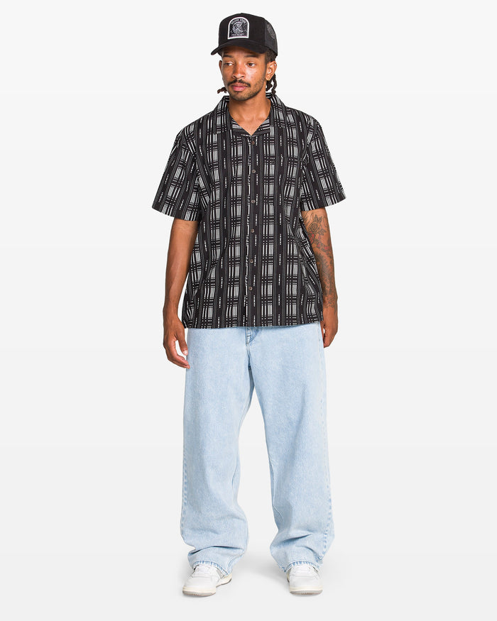 A man stands against a plain white background wearing a black patterned short-sleeve shirt, Volcom Men's Billow Denim Pant in light blue, white sneakers, and a black cap.