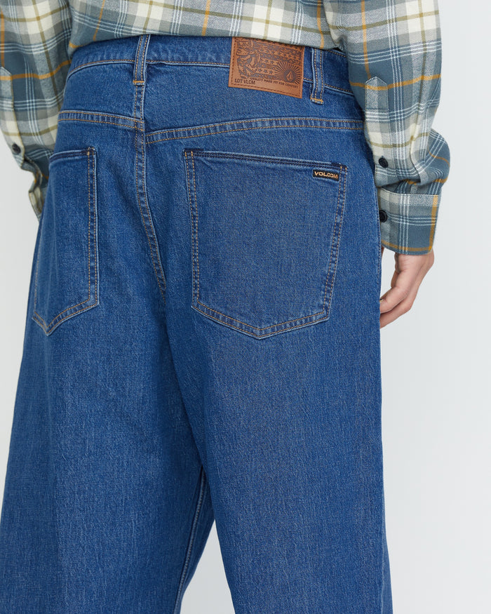 A person wears Volcom Men's Billow Denim Pants, loose fit blue jeans with a brown leather patch on the waistband and a visible Volcom label on the back pocket, paired with a plaid shirt. The image shows the back view of these low-rise jeans.