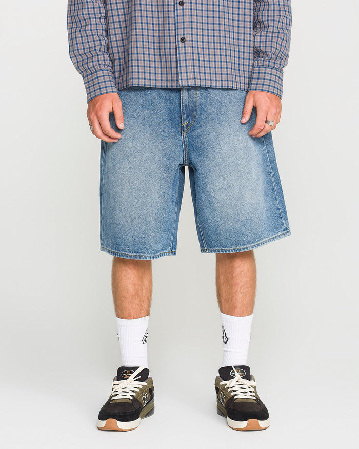 A person wearing a blue plaid shirt, loose fit Men's Billow Denim Shorts by Volcom, white socks, and black sneakers stands against a plain background.