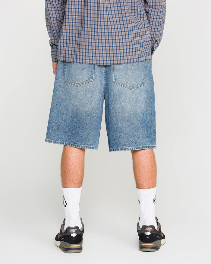 A person wearing a blue plaid shirt, Volcom Men's Billow Denim Shorts in a loose fit, white socks, and black sneakers is standing with their back to the camera against a plain background.