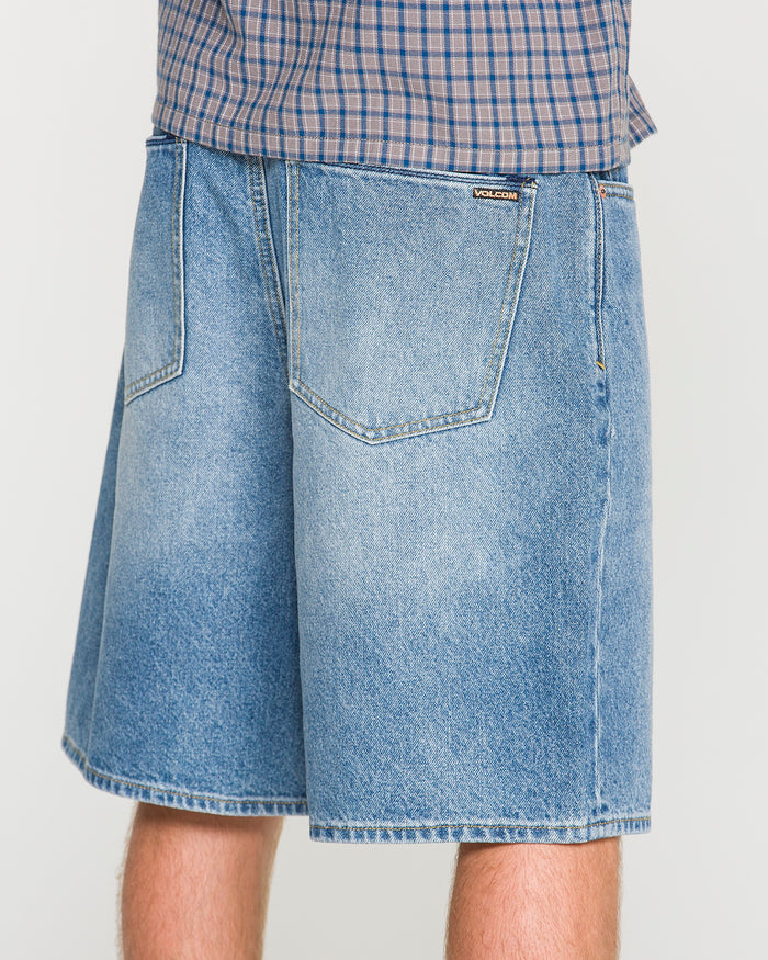 A person wearing loose fit, light blue Men's Billow Denim Shorts by Volcom and a plaid shirt, shown from the waist down, facing away from the camera.