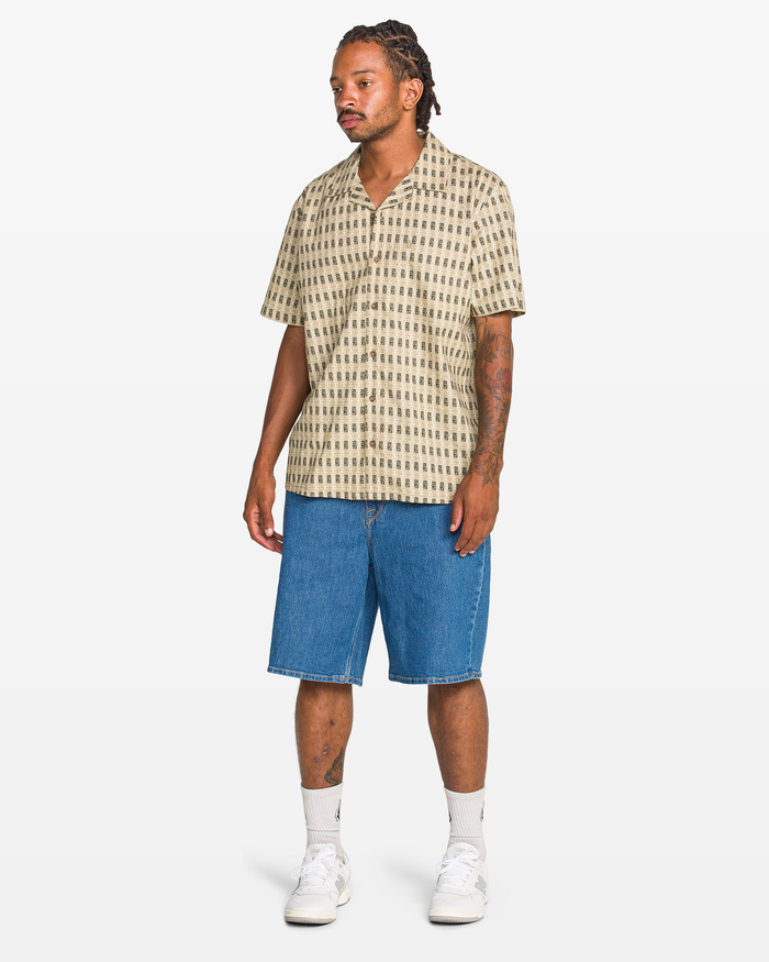 A man stands against a white background wearing Volcom Men's Billow Denim Short 22, a patterned beige short-sleeve shirt, white socks, and white sneakers.