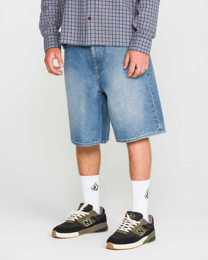 A person wears loose fit knee-length Men's Billow Denim Shorts by Volcom, along with a blue plaid shirt, white socks with a logo, and black New Balance sneakers. Only their lower body is visible.