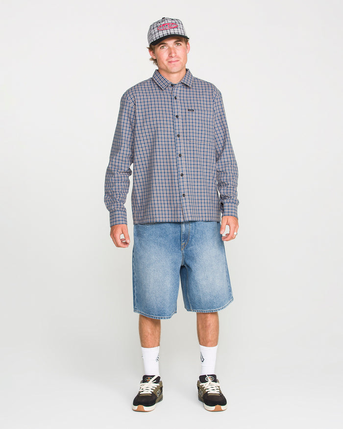 A person wearing a loose fit plaid shirt, Volcom Men's Billow Denim Shorts, sneakers, white socks, and a baseball cap stands against a plain white background.