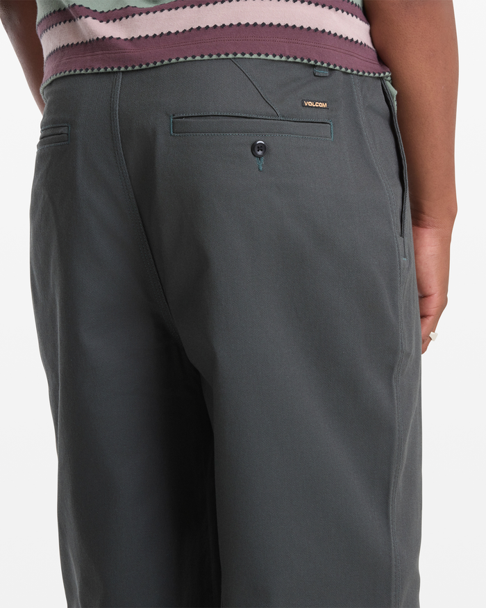 A person stands with their back to the camera, wearing Volcom Men's Billow Pants in dark gray. The pants feature a buttoned back pocket and a small gold Volcom label above the pocket.