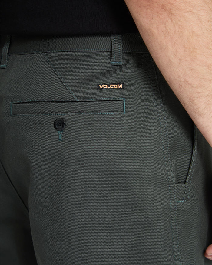 Close-up of a person wearing Volcom Men's Billow Pants in dark green, highlighting the back pocket with a button, a belt loop, and the Volcom logo tag. Part of their arm and black shirt are also visible.