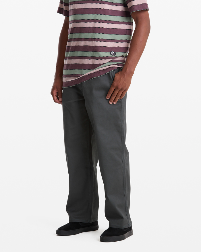 A person models Volcom Men's Billow Pants—loose-fitting and gray—with a short-sleeved striped shirt and black slip-on shoes. The individual's head is not visible, and the background is plain white.