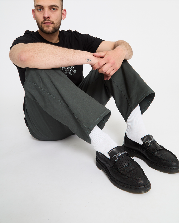 A man with short hair and a beard sits on the floor against a white background, wearing a black t-shirt, Volcom Men's Billow Pants in dark green, white socks, and black loafers.