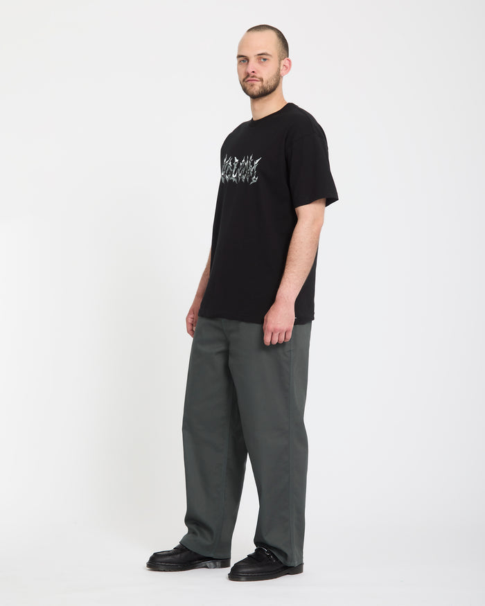 A bearded man stands against a plain white background, wearing a black graphic T-shirt, black shoes, and Volcom Men's Billow Pants in dark gray. He looks slightly to the side with a neutral expression.