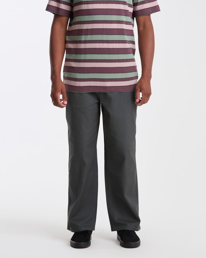 A person, head not visible, models Volcom Men's Billow Pants paired with a short-sleeve striped shirt and black shoes on a plain white background.