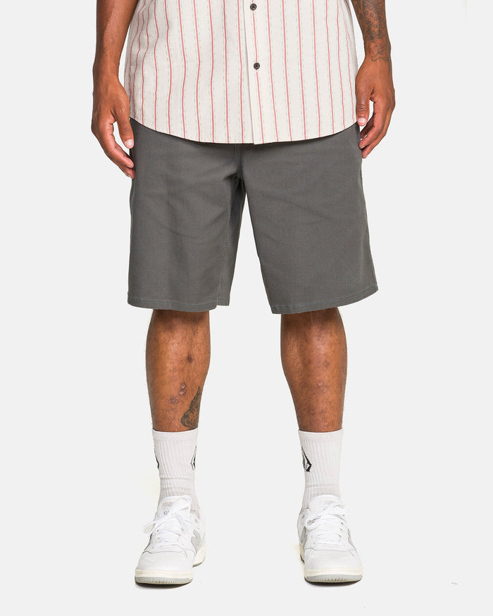 A person in a white short-sleeve, button-up shirt with red stripes, Volcom Men's Billow Shorts in castlerock twill, white socks, and white sneakers stands against a plain white background.