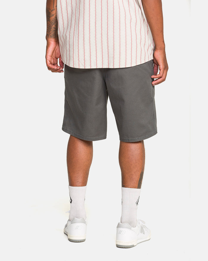 A person, shown from the waist down against a white background, wears Volcom Men's Billow Shorts in castlerock twill with white socks and sneakers, facing away from the camera.