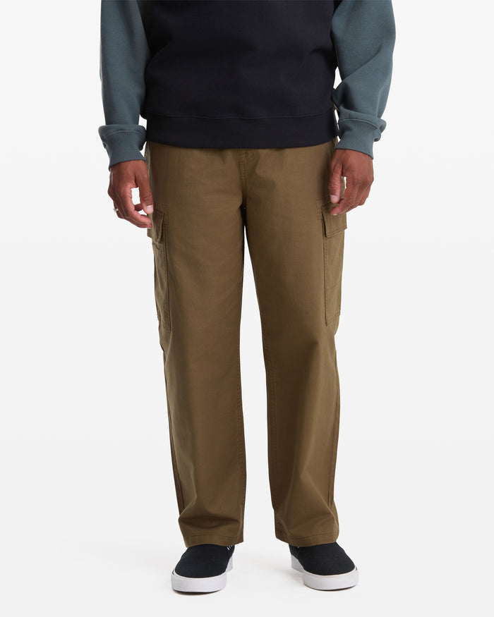 A man in Volcom Men's Billow Tapered Cargo Pants with an elastic waist, a dark long-sleeve shirt with lighter sleeves, and black slip-on shoes stands facing forward against a plain white background.