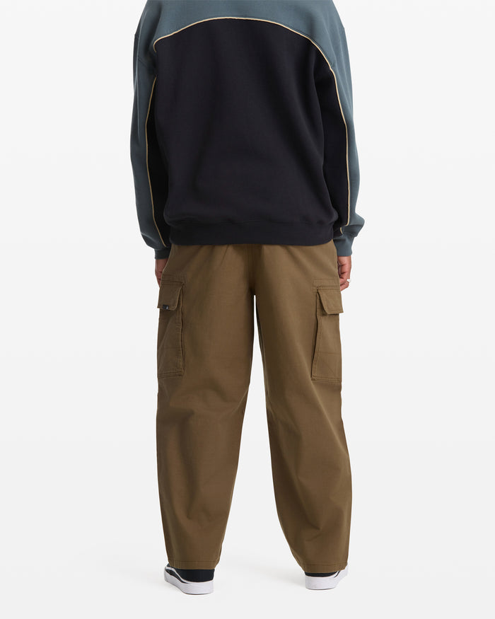 A person stands facing away, wearing Volcom Men's Billow Tapered Cargo Pants—loose-fitting cotton twill cargo pants with side pockets and an elastic waist—paired with a black and teal jacket and black-and-white sneakers, against a plain white background.