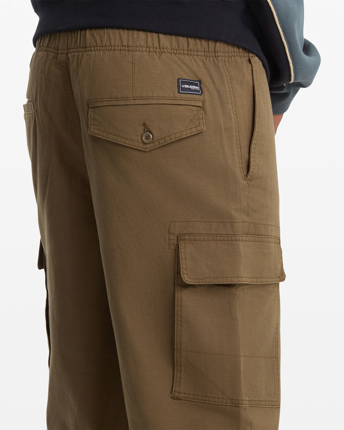 A person is wearing Volcom Men's Billow Tapered Cargo Pants featuring brown cotton twill fabric, multiple pockets including a buttoned flap pocket, side cargo pocket, and a small Volcom label on the back. Part of a dark sweatshirt is also visible.