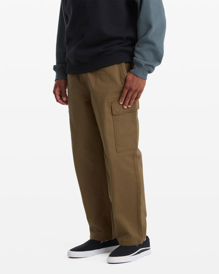 A person wears Volcom Men's Billow Tapered Cargo Pants with an elastic waist, paired with a dark sweatshirt featuring blue sleeves and black slip-on shoes, standing against a plain white background.