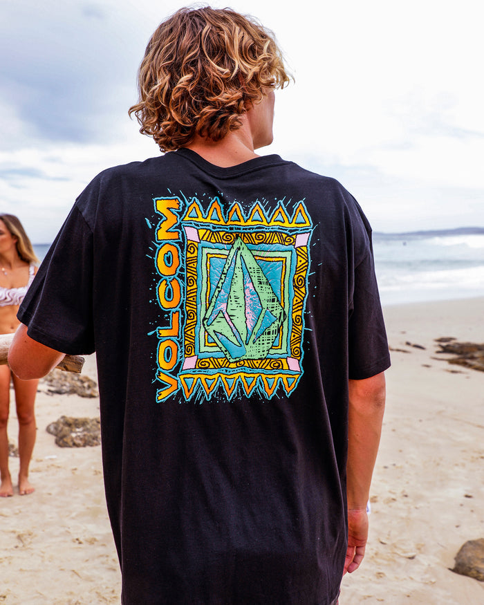 A person with wavy blond hair stands on a sandy beach, wearing the Volcom Men's Bodie Short Sleeve Tee—black with a colorful geometric back design. Another person in a bikini appears in the background by the shore.