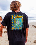 A person with wavy blond hair stands on a sandy beach, wearing the Volcom Men's Bodie Short Sleeve Tee—black with a colorful geometric back design. Another person in a bikini appears in the background by the shore.