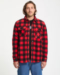 A man stands facing forward against a white background, wearing the Volcom Men's Bowered Fleece Long Sleeve in red and black plaid over a black t-shirt and blue jeans.