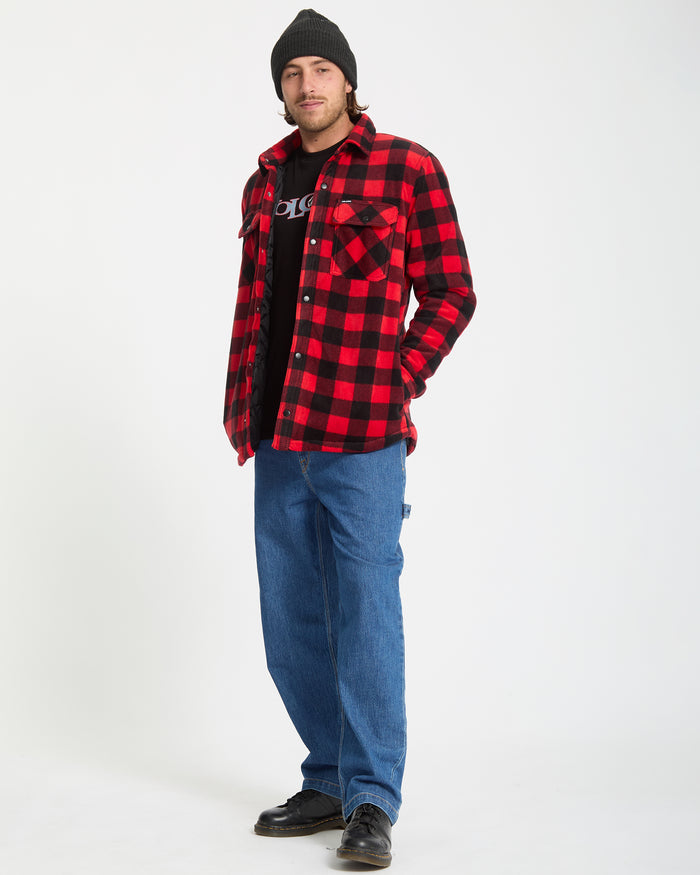 A person stands against a white background wearing a black beanie, Volcom Men's Bowered Fleece Long Sleeve in red and black plaid over a black T-shirt, loose blue jeans, and black shoes.