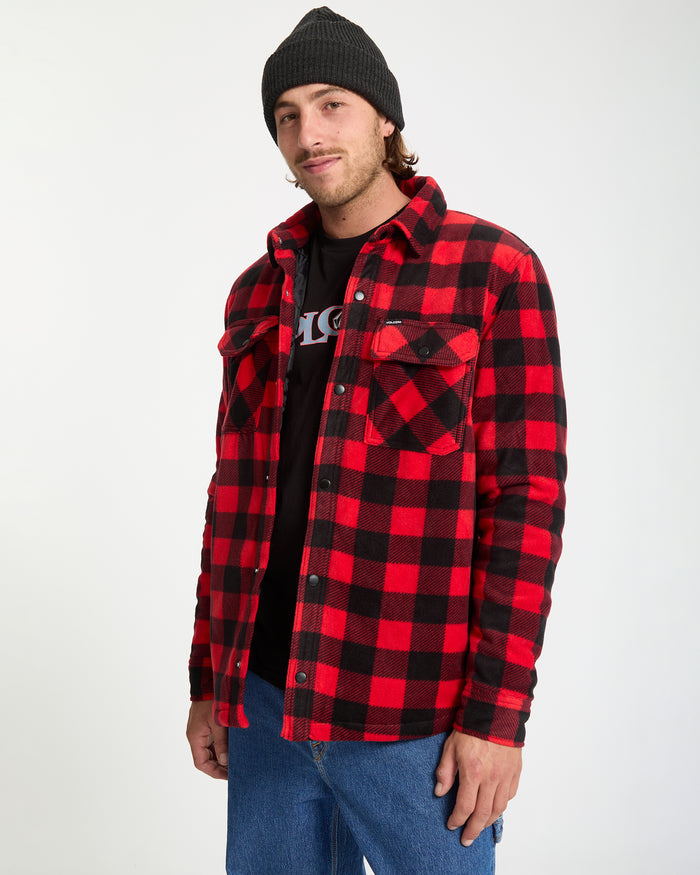 A man wearing a black beanie, Volcom graphic t-shirt, Men's Bowered Fleece Long Sleeve by Volcom in red and black plaid, and blue jeans stands against a white background, looking at the camera with a slight smile.