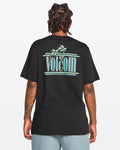 A person with braided hair, seen from behind, wears a Volcom Men's Breathe Deep Short Sleeve Tee in black with a turquoise palm tree logo, plus light gray shorts and a left arm tattoo.