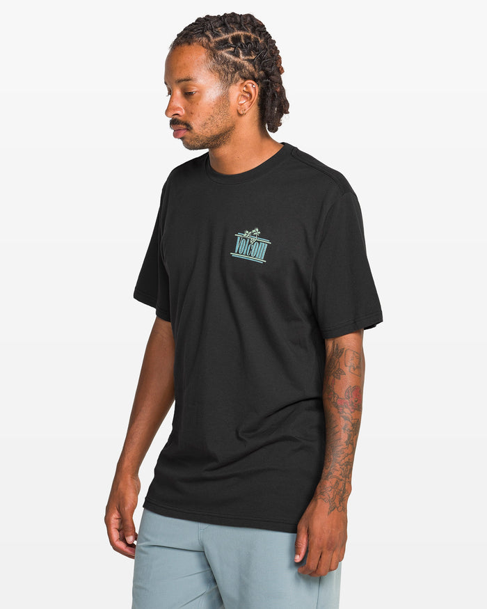 A man with braided hair and arm tattoos wears a Volcom Men's Breathe Deep Short Sleeve Tee featuring a small blue chest graphic and light blue shorts, standing against a plain white background.