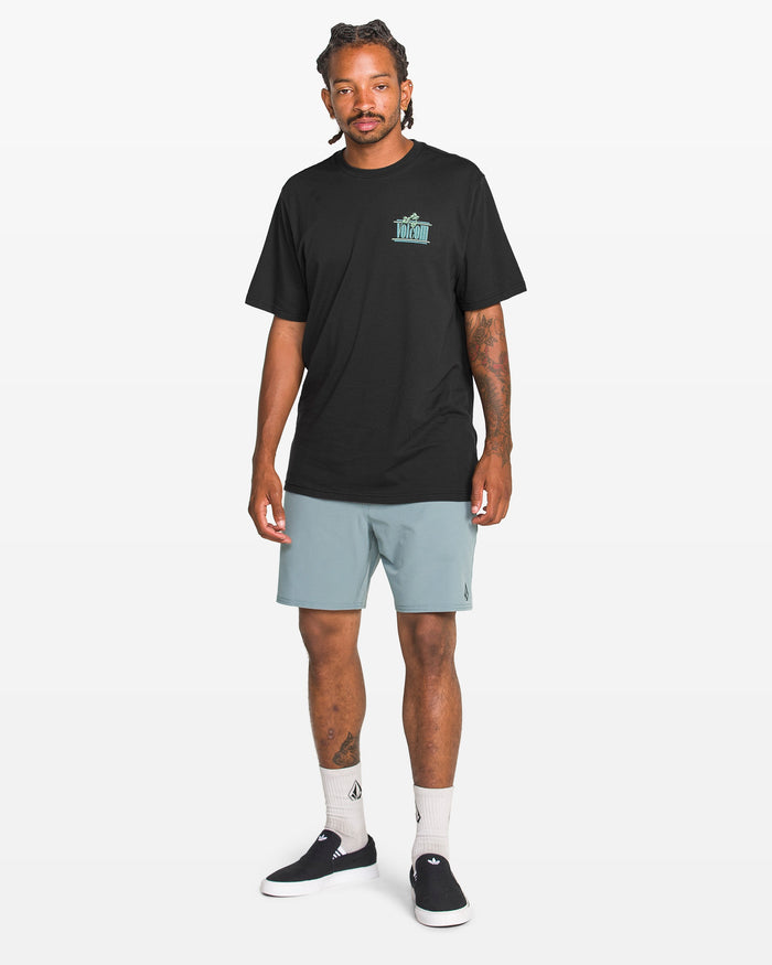 A man with braided hair stands against a white background wearing the Volcom Men's Breathe Deep Short Sleeve Tee in black with a small teal graphic, light blue shorts, white socks, and black slip-on sneakers.