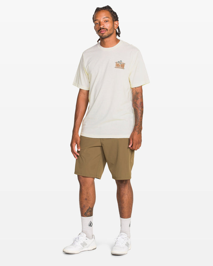 A man with braided hair and tattoos wears the Volcom Men's Breathe Deep Short Sleeve Tee featuring a chest graphic, paired with brown shorts, white socks, and white sneakers against a plain background.