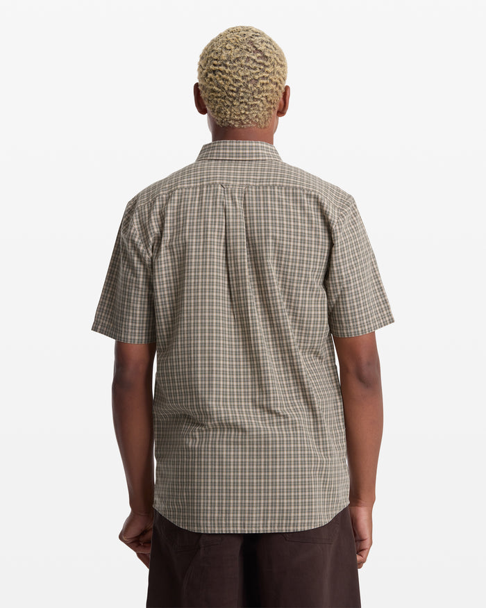 A person with short, light blonde hair is shown from the back wearing the Volcom Men's Brewster Short Sleeve shirt in brown and beige plaid, paired with dark pants, standing against a plain white background.