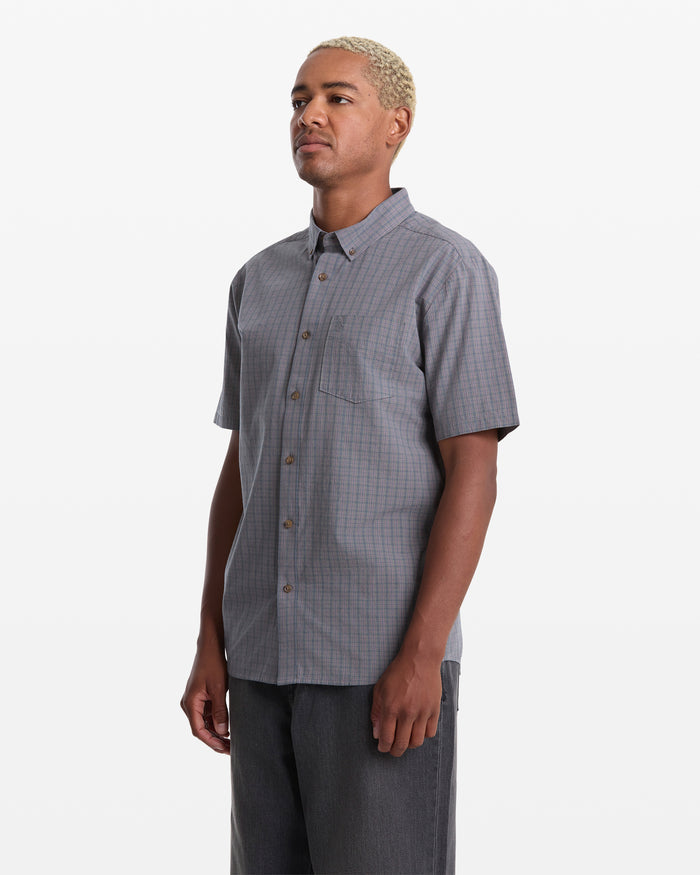 A person with short blond hair wears the Volcom Men's Brewster Short Sleeve shirt, featuring a fine plaid pattern, front pocket, and gray color, standing against a plain white background.