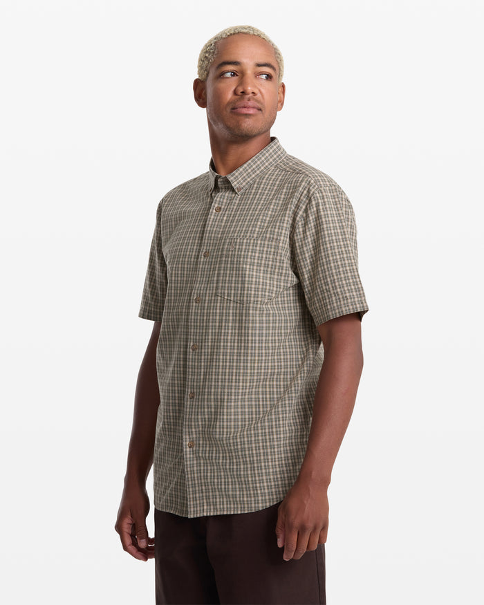 A person with short, light blond hair models the Volcom Men's Brewster Short Sleeve shirt with dark pants, standing and looking slightly to the side against a plain white background.