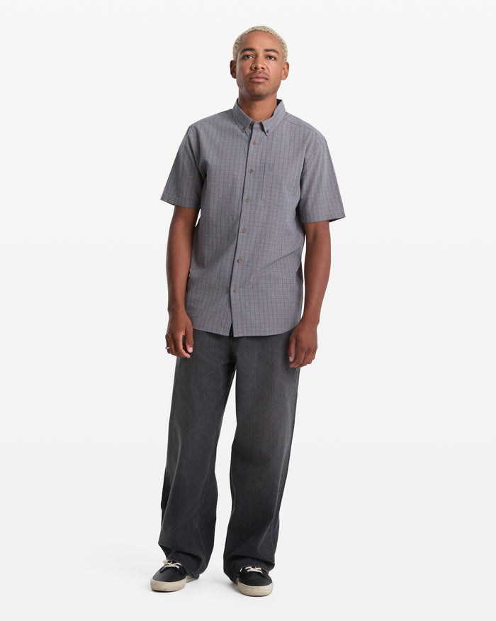A person with short blond hair wears the Volcom Men's Brewster Short Sleeve shirt, wide-leg dark jeans, and black sneakers, standing against a plain white background.