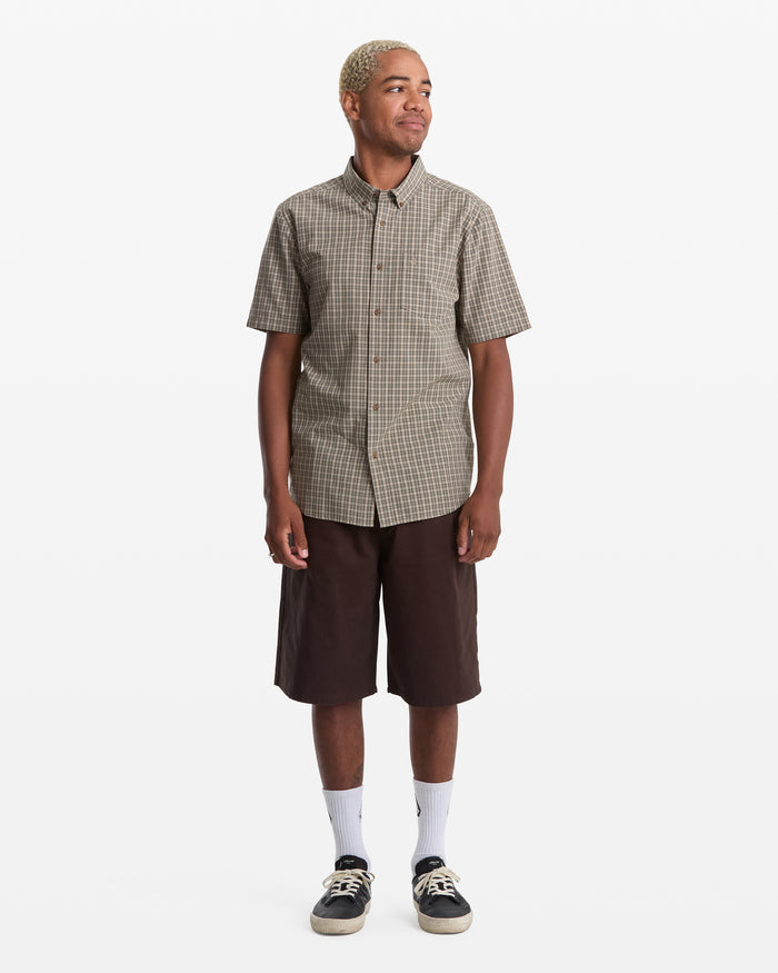 A person with short blonde hair wears the Volcom Men's Brewster Short Sleeve shirt, brown knee-length shorts, white crew socks, and black sneakers, standing against a plain white background.
